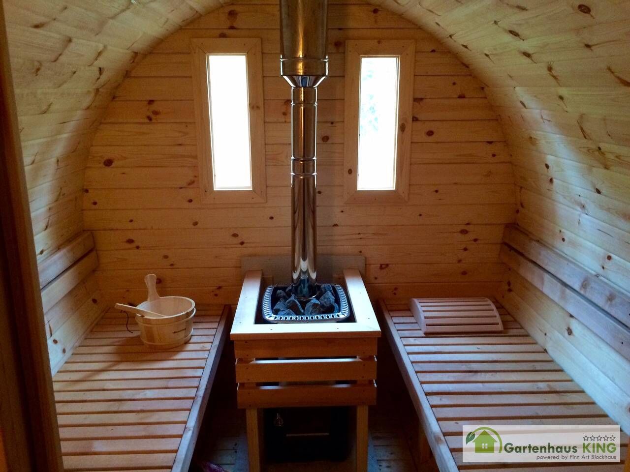 Finn Art Fass-Sauna Premium Innenansicht, Holzofen mit Dachdurchführung vor Rückwand mit zwei Fenstern, Bänke, Kübel, Aufgusskelle und Kopfstütze